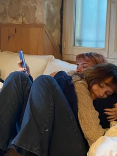 two women sitting on a bed with their arms around each other and one holding a cell phone