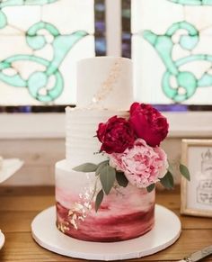 a three tiered cake with flowers on top sitting on a wooden table next to a stained glass window