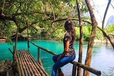 a woman is sitting on a wooden bridge over the blue water and looking at the trees