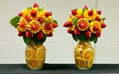 two vases filled with fruit and flowers on top of a black mat in front of a white wall