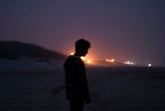 a man standing in the snow at night with his back turned to the camera and lights behind him