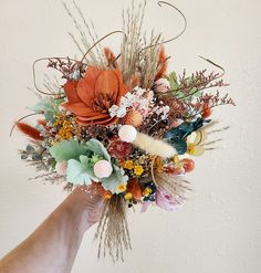 a hand holding a bouquet of dried flowers and grasses in front of a white wall