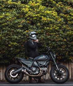 a person standing next to a motorcycle on the street with bushes in the back ground