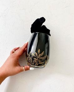 a hand holding a black and gold vase with flowers on it's side against a white wall