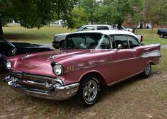 an old pink car parked next to other cars in a field with trees and grass