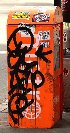 an orange box with graffiti on it sitting next to a pole and some trash cans