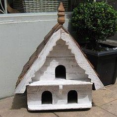 a white bird house sitting on the ground next to a potted plant in front of a building