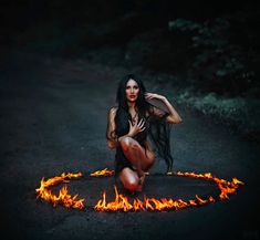 a woman sitting on the ground in front of fire with her hands behind her head