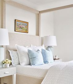 a bed with white sheets and blue pillows in a bedroom next to a painting on the wall