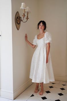 a woman in a white dress standing next to a wall with a clock on it