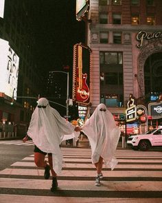 two women dressed in ghost costumes cross the street at an intersection with neon signs and buildings behind them