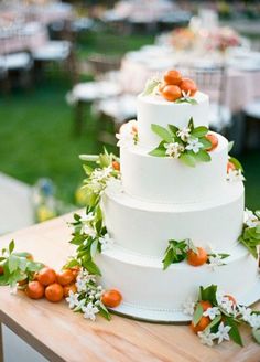 a white wedding cake with oranges and greenery on the top is sitting on a wooden table