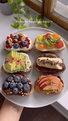 a person holding a white plate with different types of sandwiches and fruit on it in front of a window