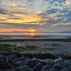 the sun is setting over an ocean with rocks and grass on the ground in front of it