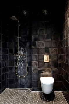 a black tiled bathroom with a white toilet and shower head on the wall, along with herringbone tile flooring