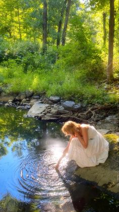 a woman in a white dress is kneeling on the bank of a river and looking at water