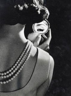 a woman wearing pearls and holding a mirror to her face