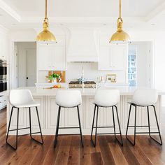 three white chairs sitting in front of a kitchen island with gold pendant lights over it