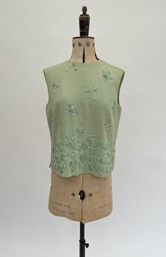 "This very pretty late 1950s to early 1960s vintage sleeveless top or blouse is in a lovely shade of pale green and is decorated with hand sewn beaded ribbon flowers. The outer fabric is a silk chiffon with the lining made from a synthetic fabric. It opens and closes at the back with a zip. The inside label says it was made in Hong Kong exclusively for the Hetty Bradley boutique.  The inside label says size 38 but this is an old size, so please go by the measurements given here to see if it will fit you. When laid flat and fully zipped at the back it measures 18.5\" inches across from underarm to underarm across. It is 18\" inches across at the waist. When zipped, the neck opening is 6\" inches across. It is in good overall condition and has been steam cleaned before sale. On close inspect Green Sleeveless Evening Blouse, Vintage Green Silk Top, Silk Summer Tops With Floral Embroidery, Vintage Sleeveless Wedding Tops, Elegant Green Embellished Tops, Embellished Sleeveless Silk Blouse, Sleeveless Embellished Silk Blouse, Spring Embellished Silk Tops, Vintage Embellished Sleeveless Top