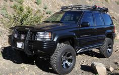 a black suv parked on the side of a mountain with rocks and bushes around it