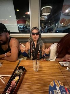 three people sitting at a table with food in front of them and one person wearing sunglasses