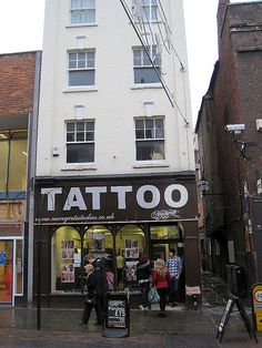 people walking in front of a tattoo shop on a city street with tall buildings behind them