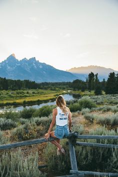 a woman leaning on a fence looking at the mountains in the distance with her back to the camera