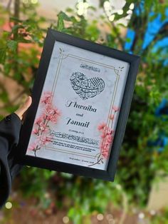 a person holding up a framed poster in front of some trees and bushes with pink flowers