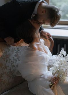 a bride and groom kissing on a couch in front of a window with white flowers