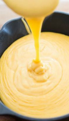 a yellow sauce being poured into a black pot filled with cheesecake batter and melted cheese