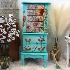 a blue dresser with flowers painted on the front and sides, sitting next to a vase filled with cotton floss