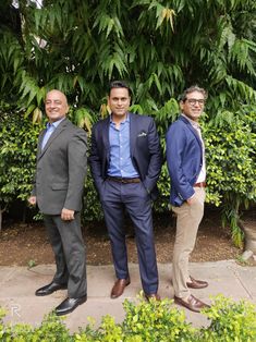 three men standing next to each other in front of some bushes and trees with green leaves