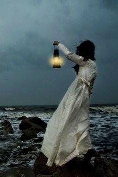 a woman in white dress holding a lantern by the ocean at night with storm clouds