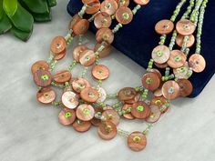 A large bib statement necklace, featuring green glass beads and orange tone mother of pearl shell disks and sequins. By Joan Rivers.  Necklace measures about 20" up to about 23" long depending on where you hook the clasp (measured at the shortest strand). Marked with "Joan Rivers" on hang tag. Some wear to the metal finish. Props shown in photos is for display purposes only, not included in this listing. As always, satisfaction is guaranteed. Thanks for shopping Vintage In Bloom! More necklaces: Bohemian Green Shell Necklace With Round Beads, Green Bohemian Shell Necklace With Round Beads, Orange Jewelry With Large Round Beads, Green Beaded Bohemian Shell Necklace, Green Bohemian Beaded Shell Necklace, Bohemian Green Beaded Shell Necklace, Adjustable Green Beaded Shell Necklace, Vintage Orange Necklace With Round Beads, Orange Agate Round Bead Necklaces