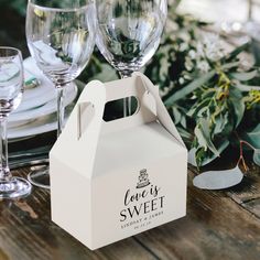 a white box sitting on top of a wooden table next to wine glasses and greenery