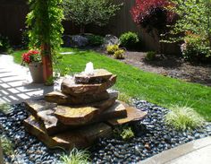 a fountain in the middle of a garden with rocks and gravel around it, surrounded by greenery