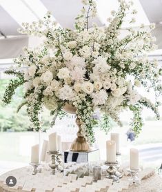 white flowers and greenery are arranged in a vase on top of a round table