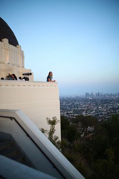 a woman sitting on top of a white building next to a tall tower with a city in the background