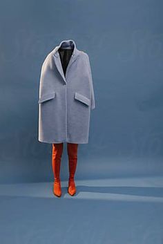 a woman standing in front of a blue background wearing an overcoat and orange boots