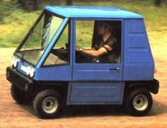 a man driving a small blue truck down a dirt road in front of a forest