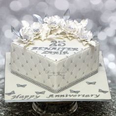 a 25th wedding anniversary cake decorated with white flowers and silver butterflies on a table in front of sparkling lights