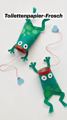 two green paper toys with eyes and legs hanging from strings on a white surface, one is shaped like a frog