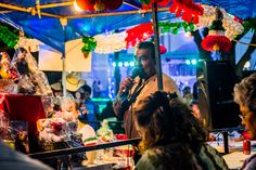 a man speaking into a microphone in front of a crowd at an outdoor market stall