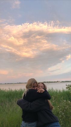 two women hugging each other in front of a body of water with clouds above them