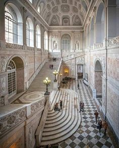 people are walking up and down the stairs in an ornate building with marble steps, chandeliers and checkered flooring