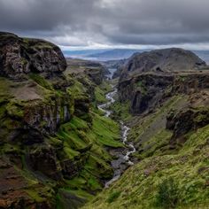 a river running through a valley surrounded by lush green grass and rocky mountains under a cloudy sky