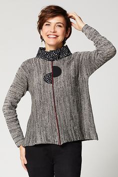 a woman in black pants and a gray sweater smiles at the camera while standing with her hands on her head