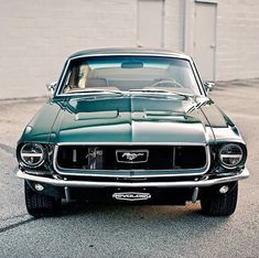 an old green mustang sitting in front of a garage door with the hood up and headlight on