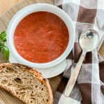 a bowl of tomato soup next to a slice of bread on a wooden cutting board