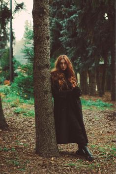 a woman standing next to a tree in the middle of a forest with long red hair